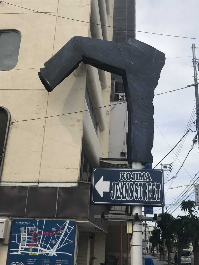児島ジーンズストリートのオブジェ。児島ジーンズストリートはこちらですというデニムの青色の看板の上にジーンズがまるで一歩踏み出しているかのようなオブジェが乗っている。