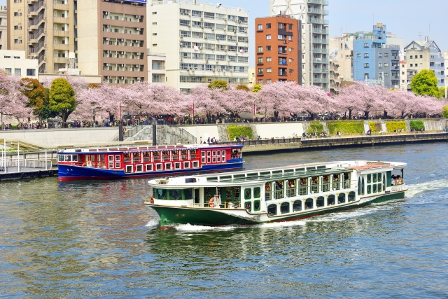 春の隅田川の写真。桜が川沿いに咲き、二隻のフェリーが隅田川を進んでいる。