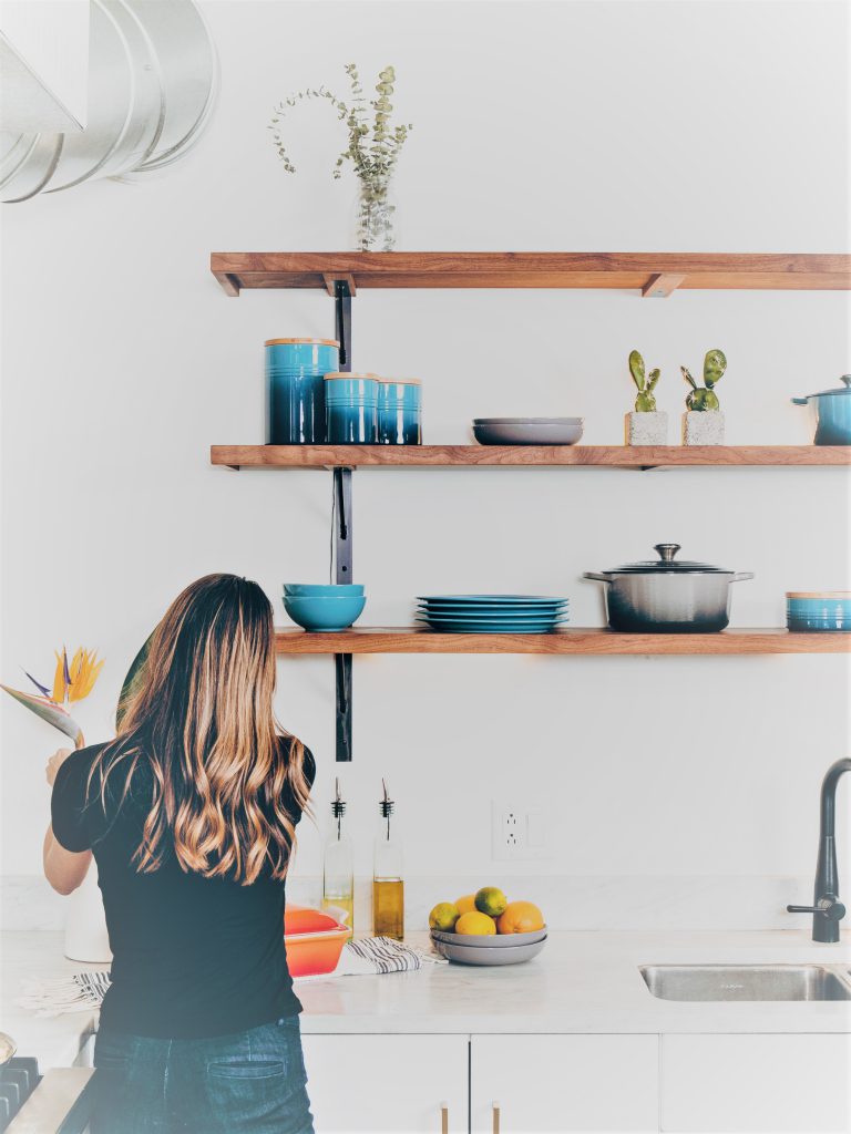 金髪の女性がキッチンに向かい調理しようとしている後ろ姿。棚にはブルーのおしゃれなホーロー鍋が置かれている。