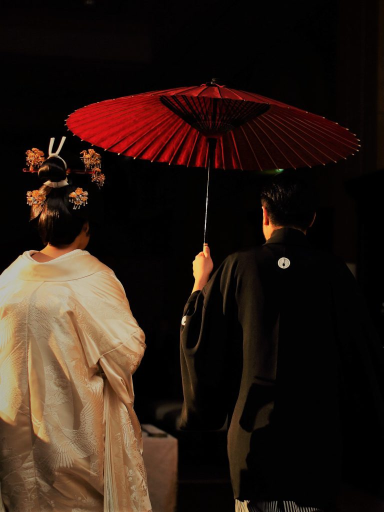 花嫁は白無垢を着て、花婿は黒い紋付き袴を着て、赤い和傘をさしながら歩いていく後ろ姿の写真。