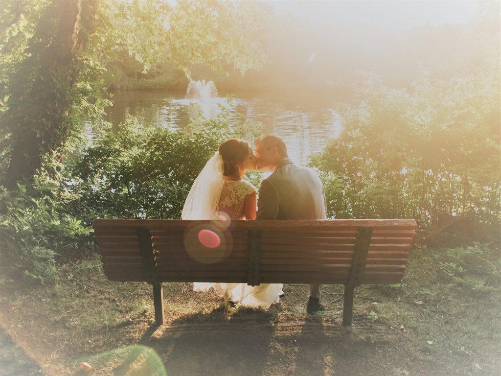 水辺のベンチに花嫁と花婿が並んで座りキスをしているところに柔らかい木漏れ日が差している