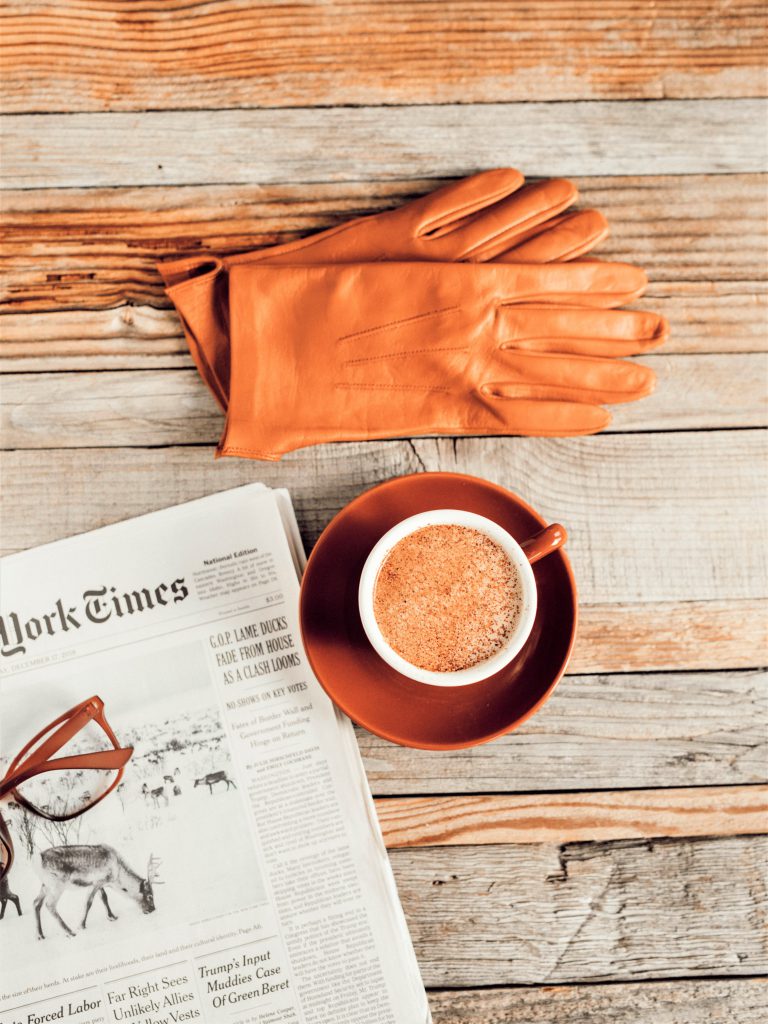テーブルの上にオレンジ色の革の手袋とコーヒーカップに入ったカプチーノと新聞と眼鏡が置いてある写真