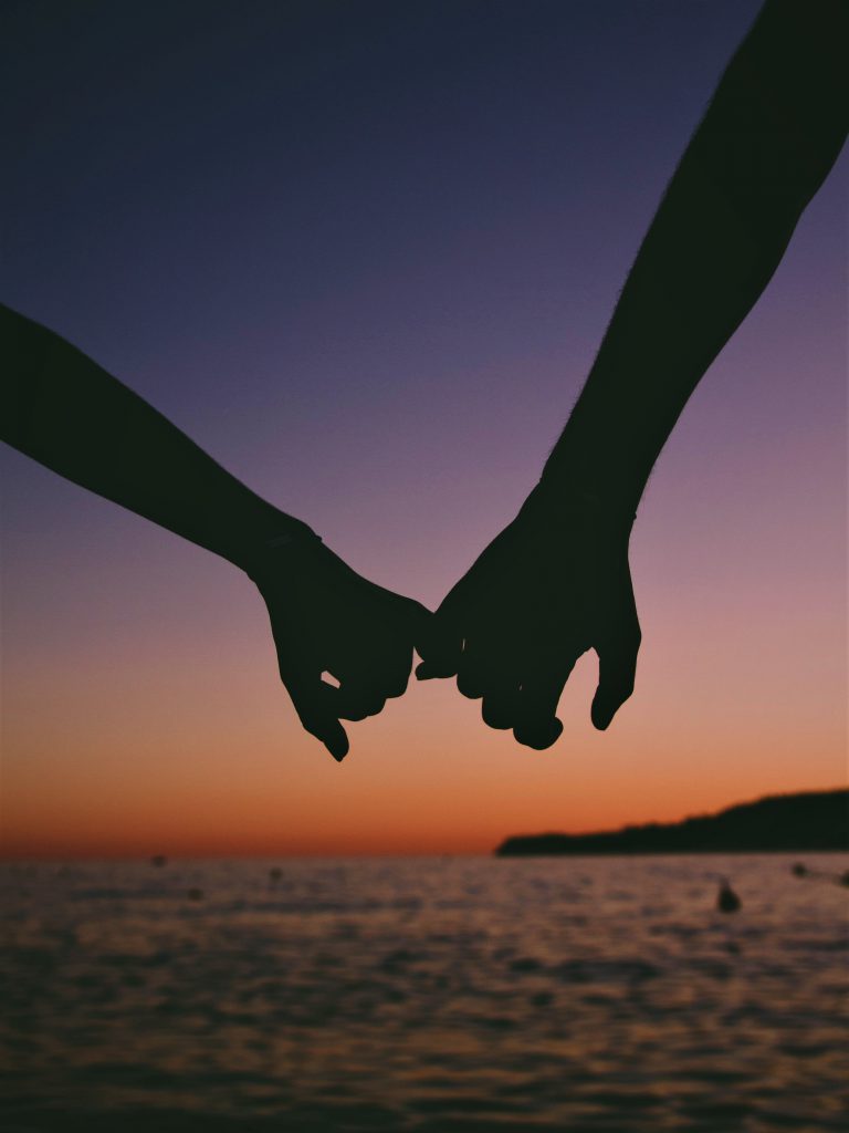 夕暮れ時の赤く染まる夕日が海に沈むのを目前にカップルが手を繋いでいる様子。