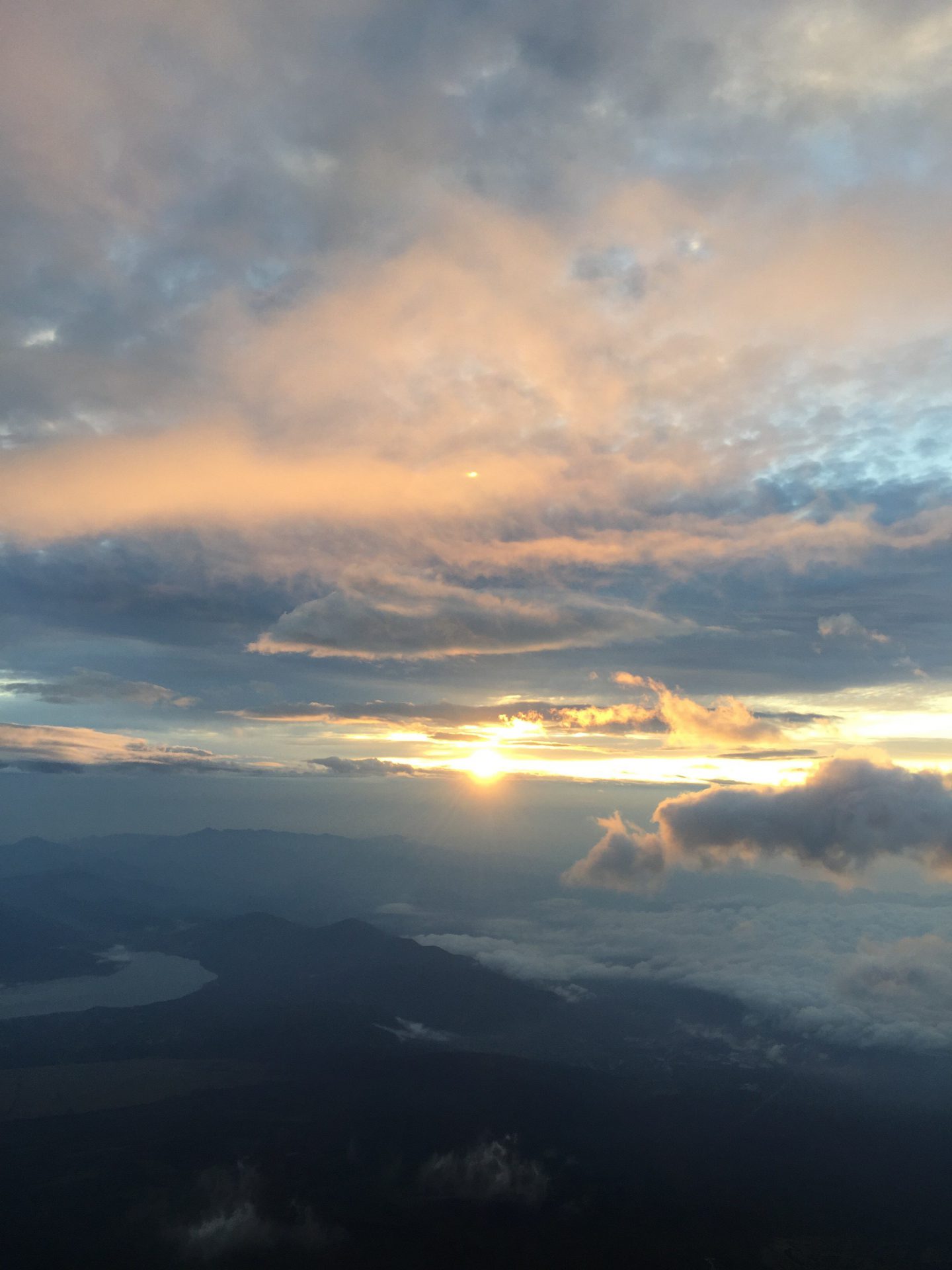 富士山ご来光（スマホ写真縦で撮影。下方に湖、はるか遠方より雲間から朝日零れる。薄いブルーからピンクへのグラデーション）