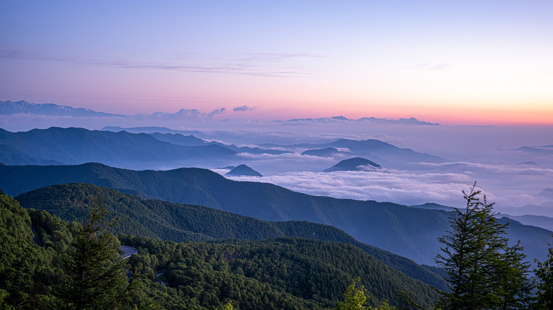 朝焼けに染まる空と雲海（闇の開ける瞬間の山々を見下ろす。はるか遠方に白い雲がたなびく姿。地平線のような空と地上との境目には影がかって濃いめの紫の雲のような色彩。その上部にうっすら赤と橙のグラデーションが浮かぶ。その向こう、靄の中に朝日の気配）