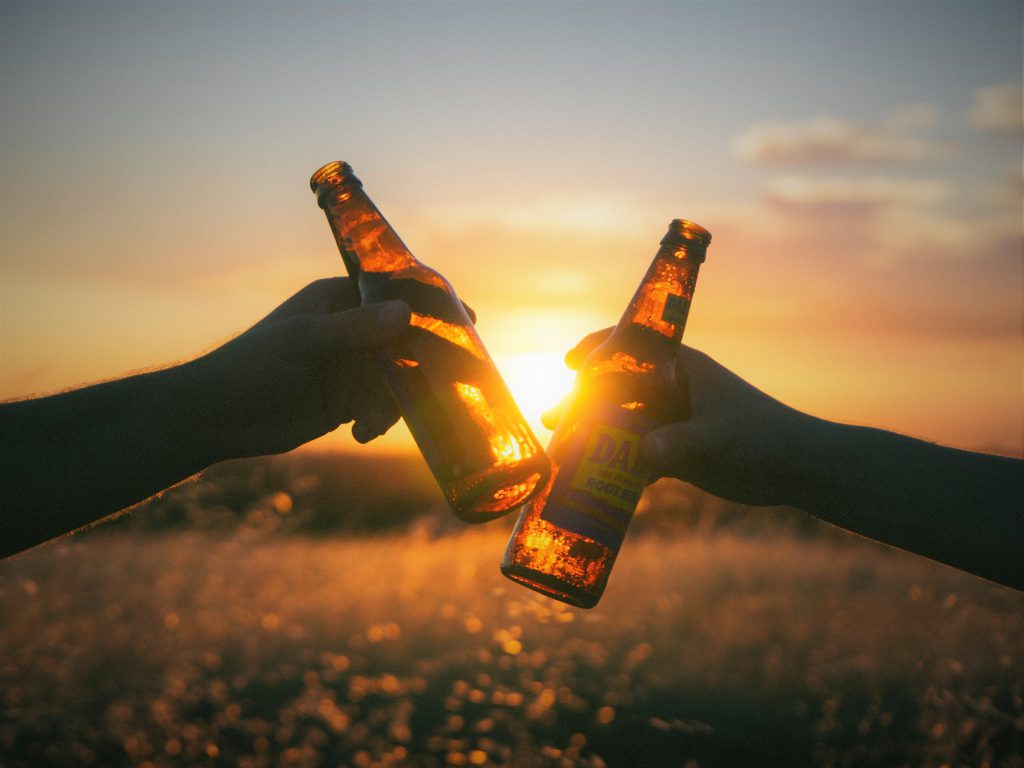 海に綺麗な夕日が沈みあたり一面はオレンジ色の光に包まれていて、幻想的な雰囲気。瓶ビールをお互いに重ね合わせて夕日に乾杯している手が2本両サイドから出ている。