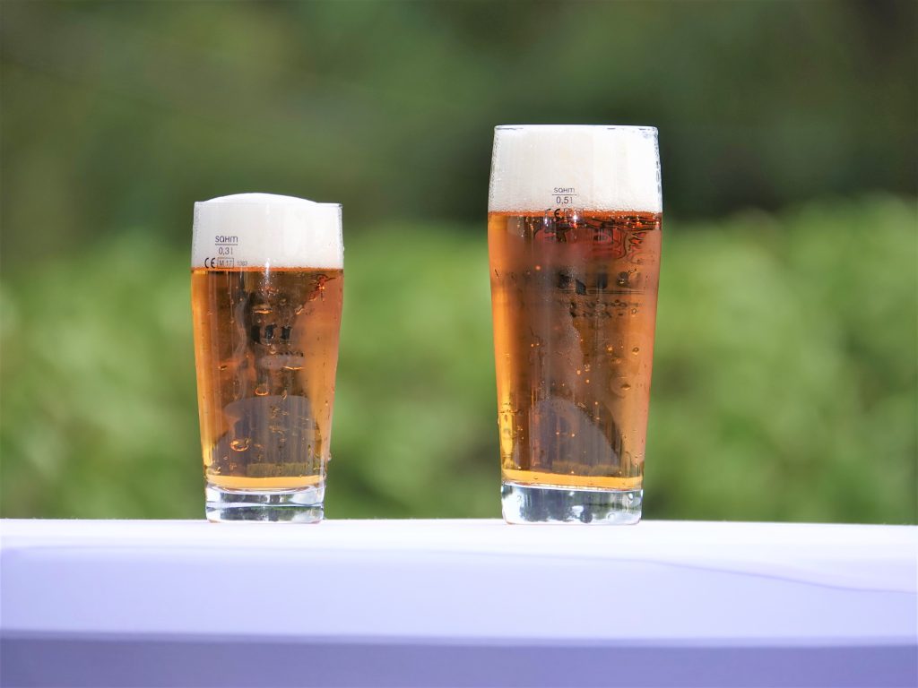 背景は緑豊かな森の雰囲気。白いテーブルに琥珀色のビールが小さいグラスと大きいグラスに注がれている。