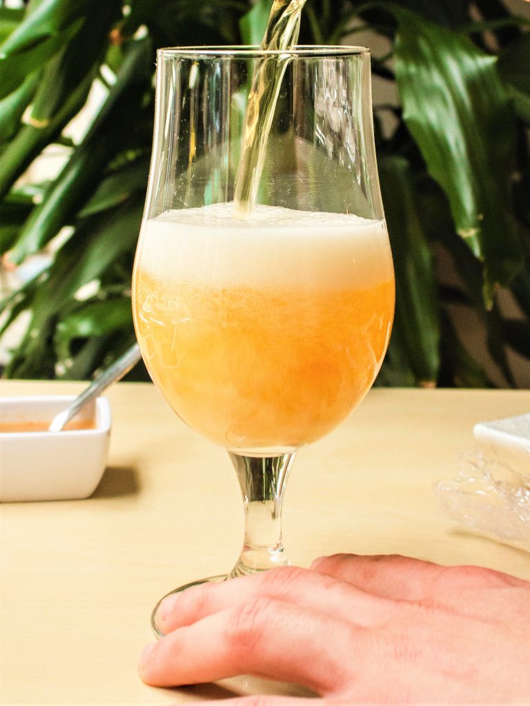 背景には緑の観葉植物があり、テーブルの上にはワイングラスに注がれている最中の黄金色のビール。グラスが動かないように手でしっかりと押さえている写真。