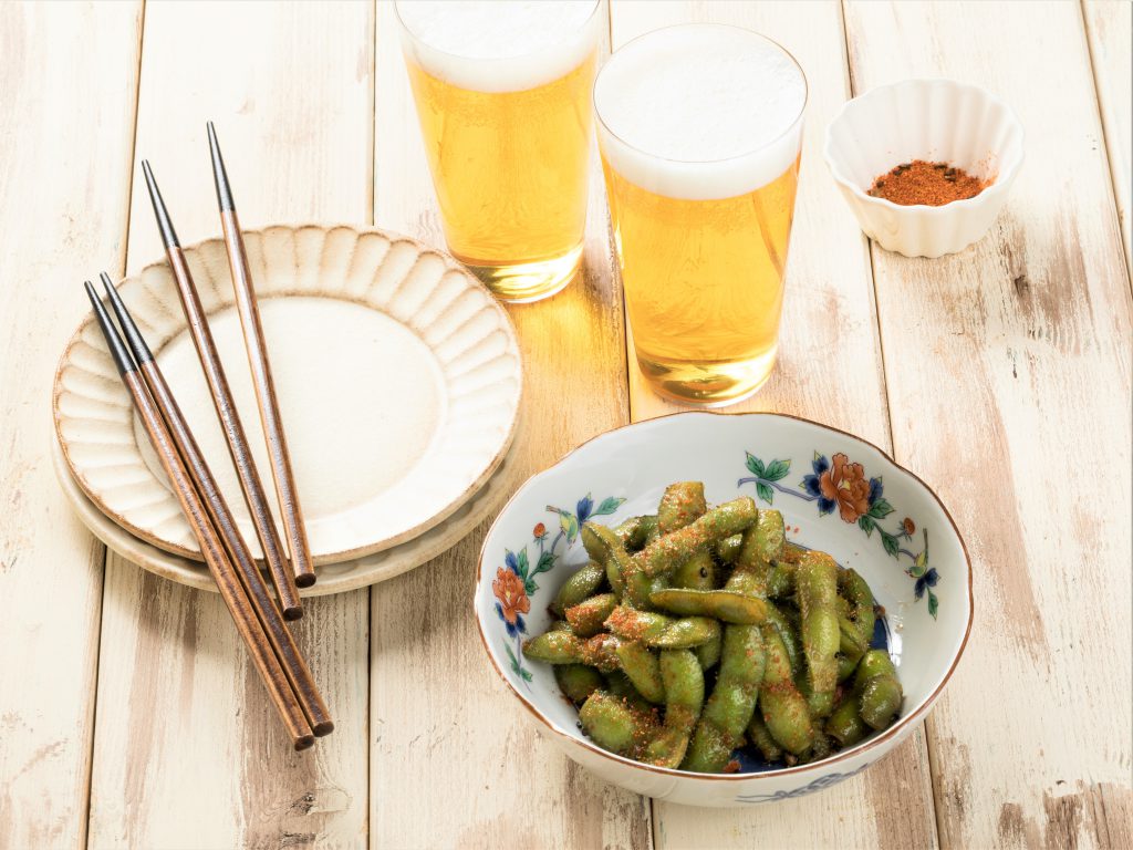 テーブルの上にビールが2杯とお皿と箸。枝豆にはスパイスのきいた赤い調味料がかけられている。