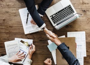 パソコンや紙資料を広げたテーブルの上に手を伸ばし、スーツを着たビジネスマンたちが握手を交わしている。