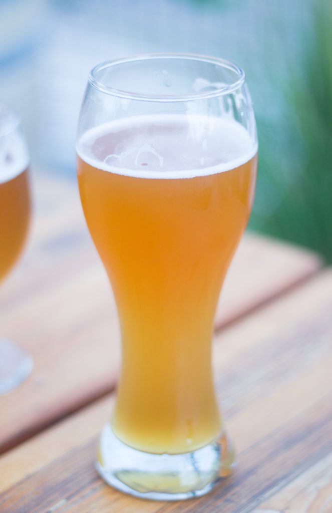 ビールグラスに注がれた白っぽい、黄金色が薄い色のビール。背後には緑っぽい湖のような景色。