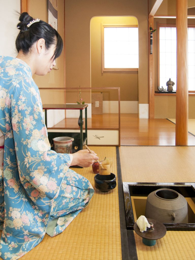 水色の着物を着た女性が畳の茶室に座ってお茶を点てている様子