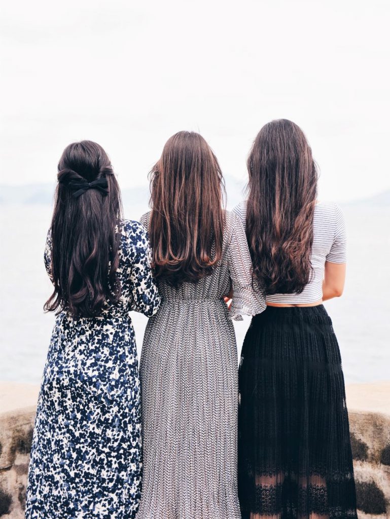 髪の長いおしゃれな服を着た女性3名が後ろ姿で海に向かって立っている
