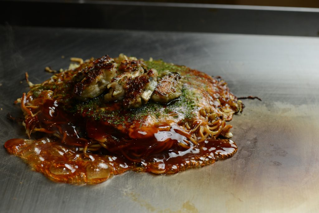 牡蠣入りお好み焼きが鉄板に乗っている様子。たれが煮立っていて美味しそう。