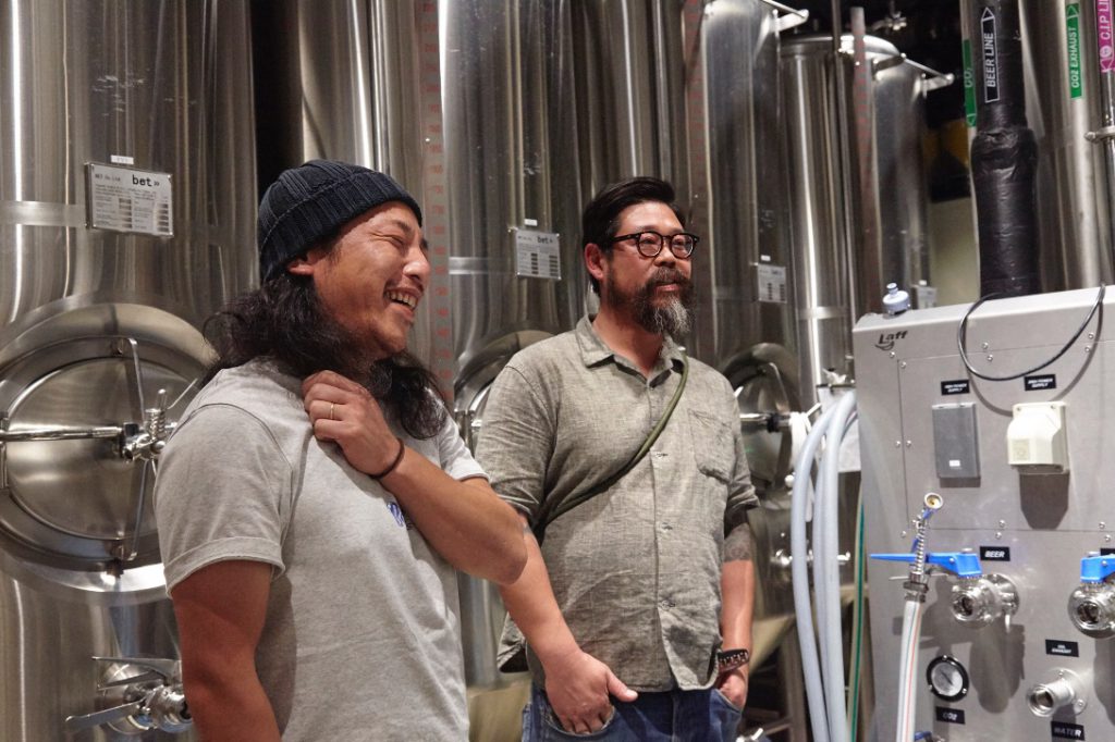 笑っている安藤氏と永石氏が銀色のビールタンクの前で立っている。