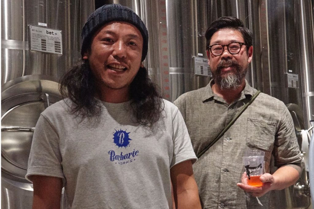 BarbaricWORKSオリジナルの、グレーのTシャツを着た安藤佑一氏と、ビールを持った永石卓宏氏が銀色のビールタンクの前で微笑んでいる。