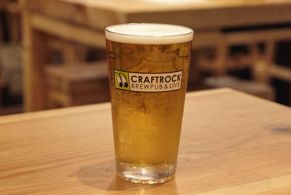 CRAFTROCKの文字の入ったグラスに注がれたCRAFTROCK BREWINGのスード ニンフ。木のテーブルに置かれている。