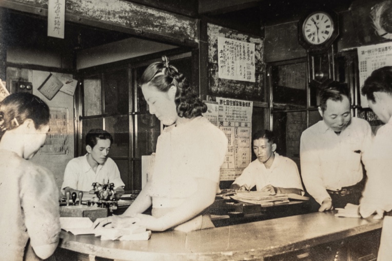 ラウンド型のカウンターで、女性が接客している様子。周りにはディスク作業している男性が見える。白黒写真で古き昭和の時代を感じる写真。