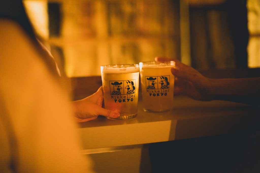 ほのかな光の中で二人の人物がMikkeller Tokyoと印字されたビールグラスを手にしており、グラスにはきめ細やかな泡が注がれています。画像の照明は柔らかく、暖かいトーンで、リラックスした雰囲気です。