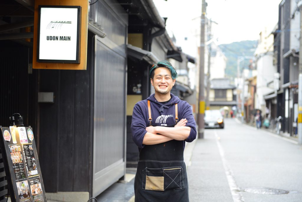 お店の入り口に立つ店主。緑のメッシュが入ったショートの髪型。濃い紫のパーカーに黒いデニム生地のエプロンをし笑顔で微笑んでいます。