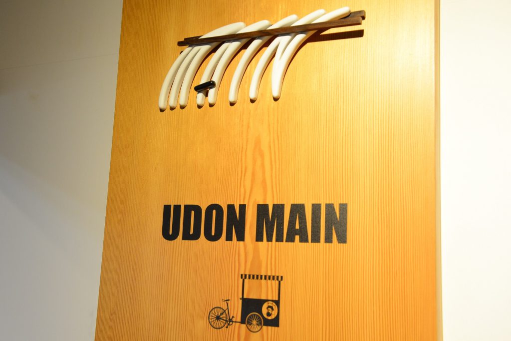 UDON MAINの看板。木の板で出来ており、上にうどんを箸でつかんだ立体的なモチーフ、真ん中にUDON MAINの文字と下に自転車を改造したリヤカーが黒色で描かれています。