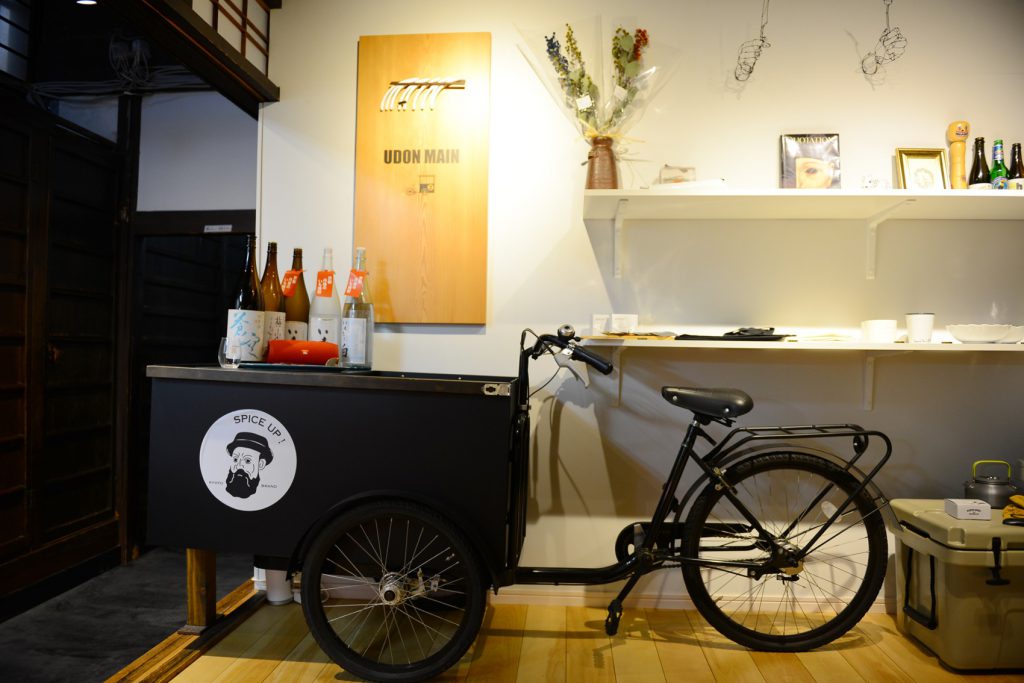 白が基調の店内に飾られた、自転車の前の部分を荷台に改造した黒色の屋台。荷台には、お酒の瓶が5本のせられています。