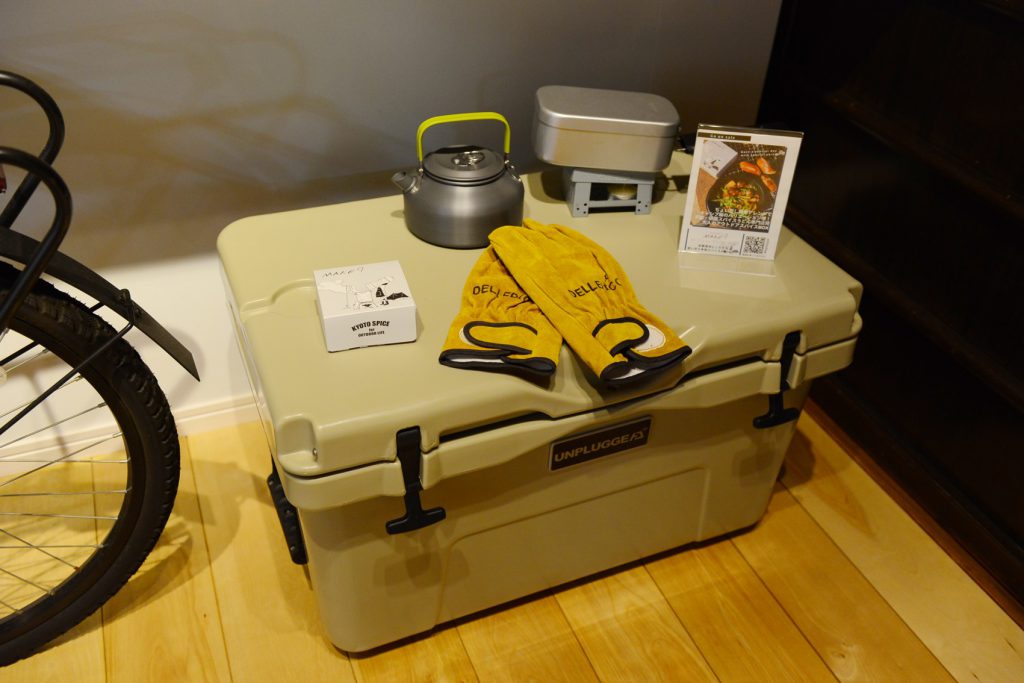 ベージュのクーラーボックスの上に、黄色い手袋、やかん、ハンゴウ、MAKE9の箱、お店のポップが置かれています。

