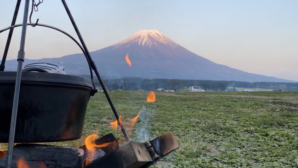 薪を使って黒い鍋で料理をしている光景。緑の平地の奥に富士山が見えます。