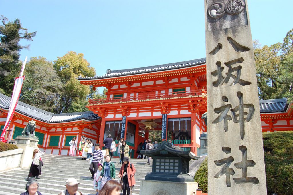 八坂神社の石碑が左側にあり、奥には朱色の神社の入り口、中央の階段は行き交う人々が見えます。