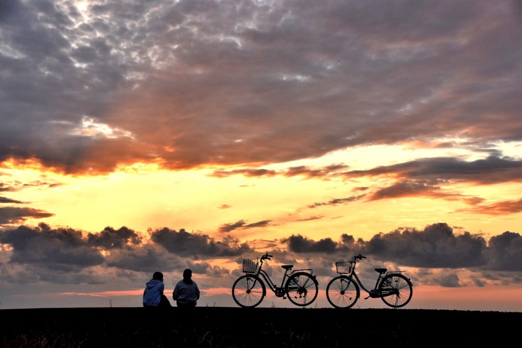自転車を止め、地面に座って美しい夕焼けを眺めている2人組。