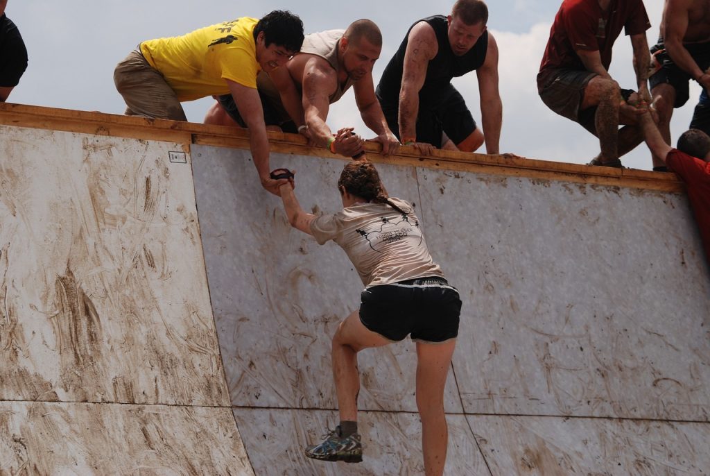 壁を登る女性の手助けをしようと壁の上から女性に手を伸ばす男性たちの様子。
