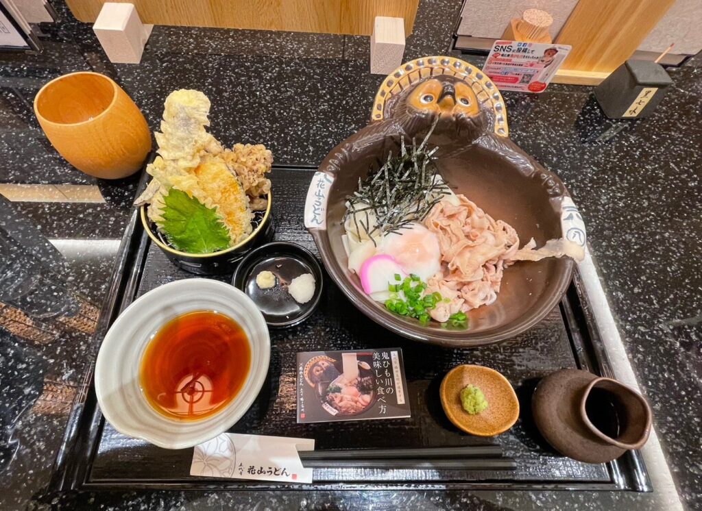群馬県の有名うどん店「花山うどん」のメニューである鬼御前。たぬきの器に幅広の郷土麺が入っており、天ぷら付き。