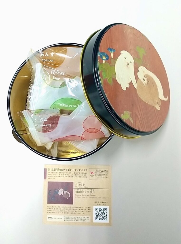 「朝顔狗子図杉戸」の絵柄がプリントされた丸い菓子缶。青い朝顔と子犬が3匹描かれている。缶の中には「彩果の宝石」という人気のフルーツゼリーが小袋でいくつか入っている。