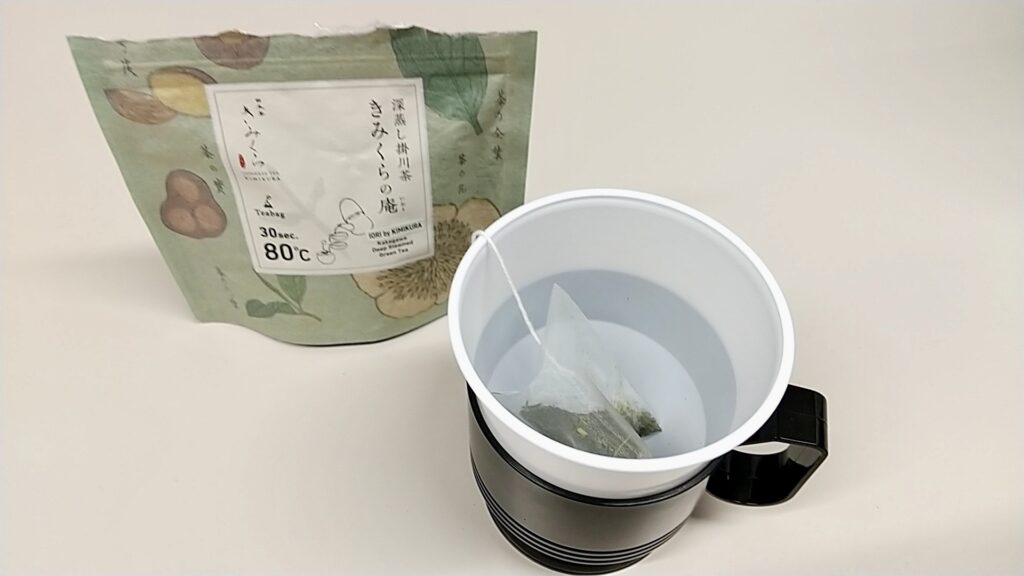 「日本茶きみくら」の商品「Art Print Package (Teabag) Collection」の1つ、羽田店限定の「深蒸し掛川茶 きみくらの庵」。緑系のパッケージの手前にはティーバッグが入ったコップが置かれている。