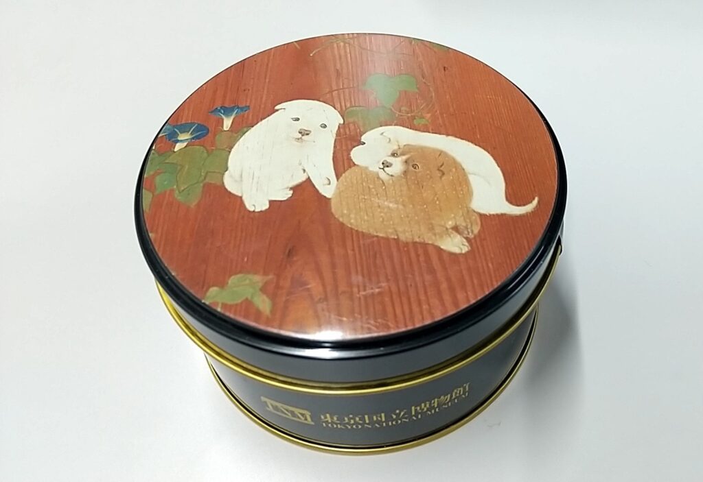 「朝顔狗子図杉戸」の絵柄がプリントされた丸い菓子缶。青い朝顔と子犬が3匹描かれている。