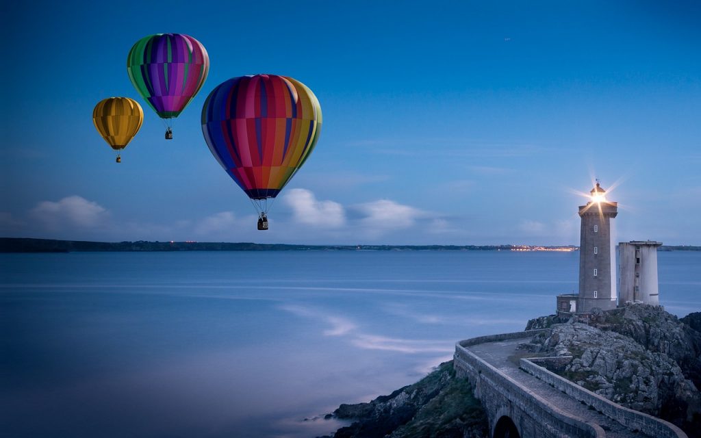 海を背景に灯台に明かりが灯り、その隣で気球が三つ浮かんでいる写真。
