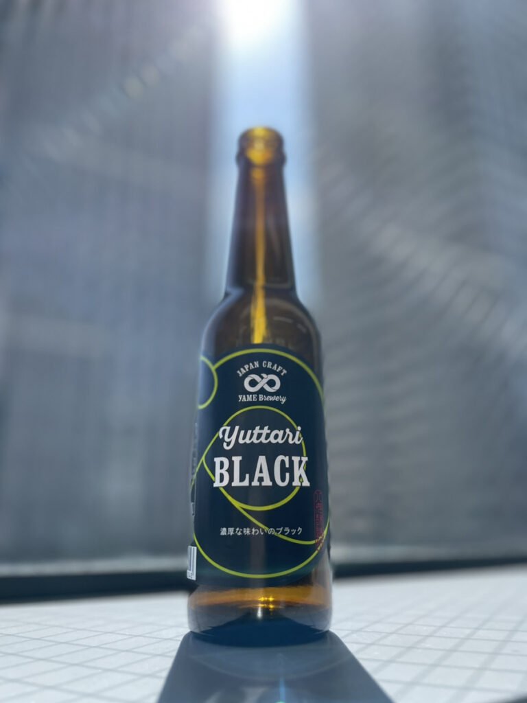 八女ブルワリーのブラック。背景にビルが見える窓辺の中央に黒色のラベルのクラフトビールの瓶が1本置いてある。