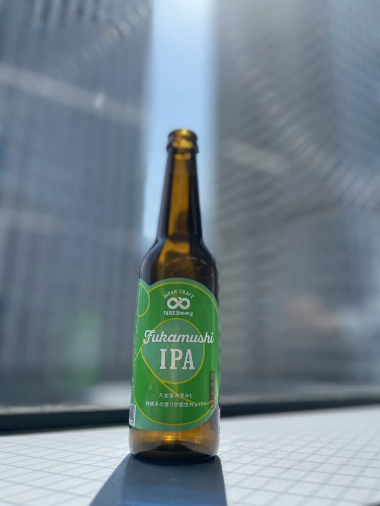 八女ブルワリーの深蒸IPA 。背景にビルが見える窓辺の中央に緑色のラベルのクラフトビールの瓶が1本置いてある。