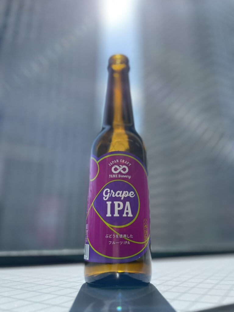 八女ブルワリーのグレープIPA。背景にビルが見える窓辺の中央に紫色のラベルのクラフトビールの瓶が1本置いてある。