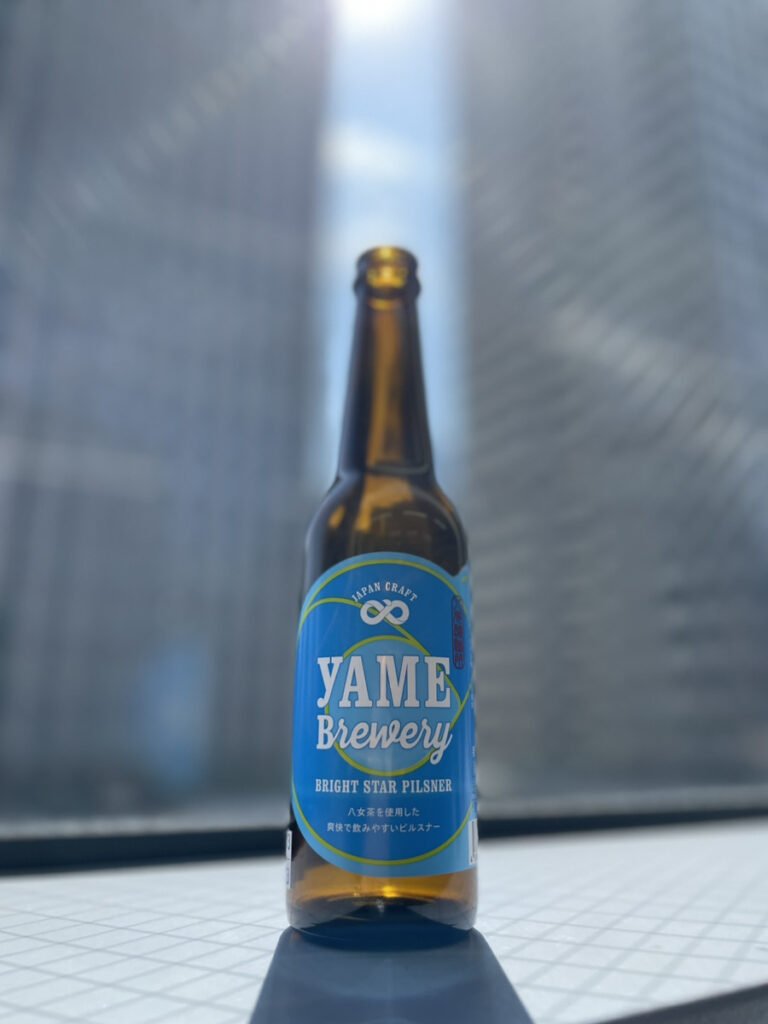 八女ブルワリーのピルスナー。背景にビルが見える窓辺の中央に青色のラベルのクラフトビールの瓶が1本置いてある。
