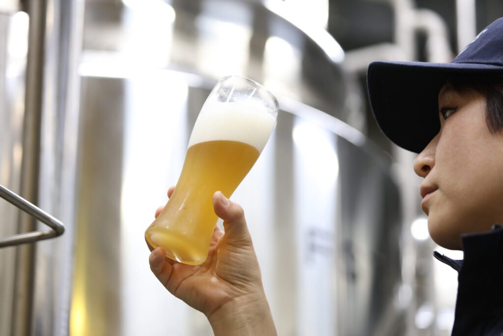 醸造所のなかで、瓶に注がれたビールを手に、見つめる紺色のキャップの帽子をかぶった醸造担当者。