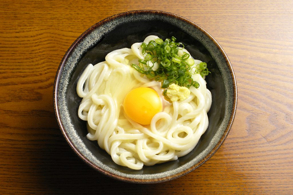 黒い器に入った窯玉うどん。生卵、生姜、ネギがトッピングされている。