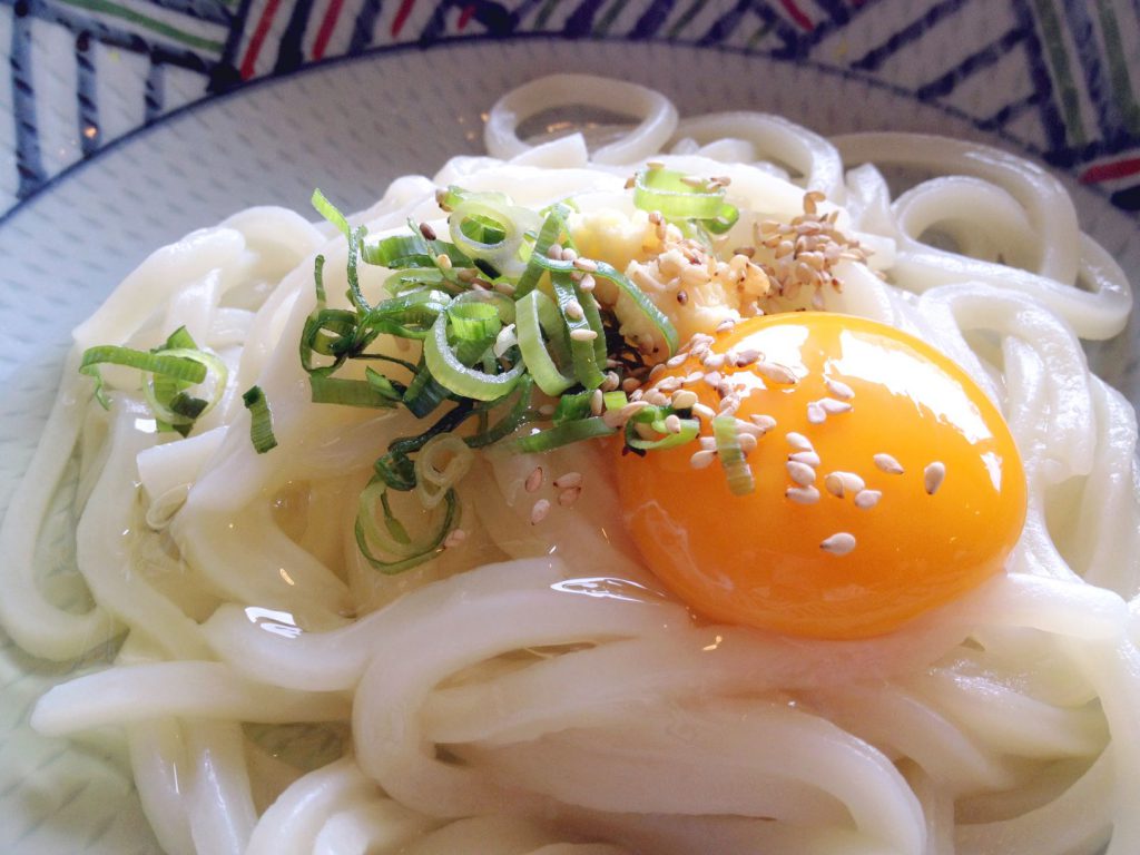 うどんの上に生卵、ネギ、しょうが、ゴマが乗っている