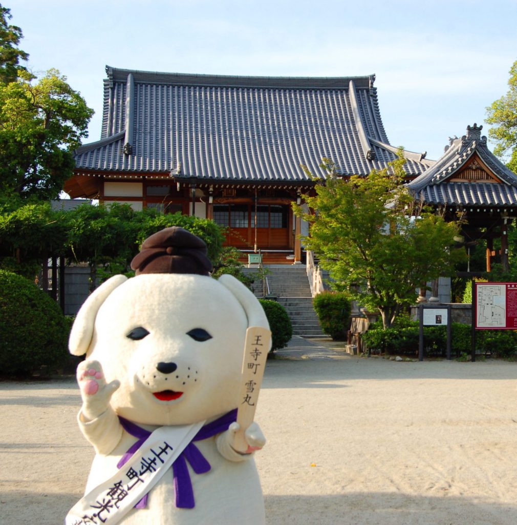 雪丸が達磨寺の前に手を挙げて立っている様子。