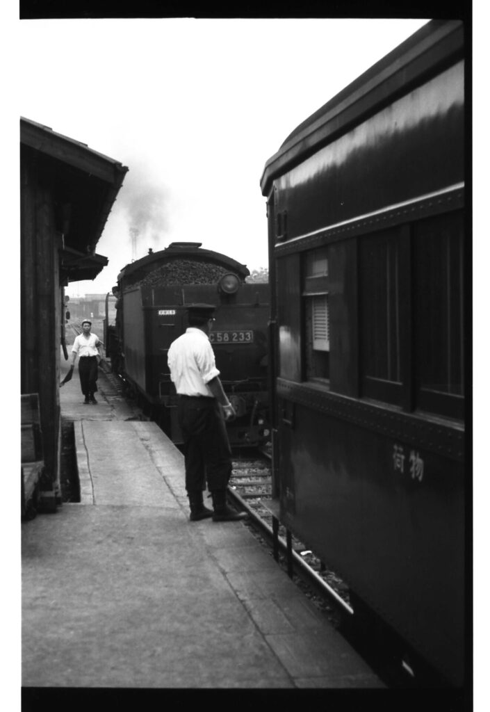 駅の職員が見守る中、荷物と表示された貨車と連結している汽車「C58　233」。石炭を積んでいるのがわかる。