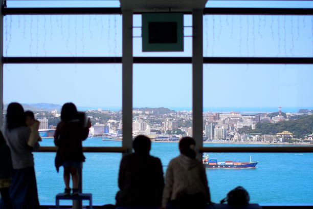 門司港レトロ展望室では人々が外の青い海を眺めている。