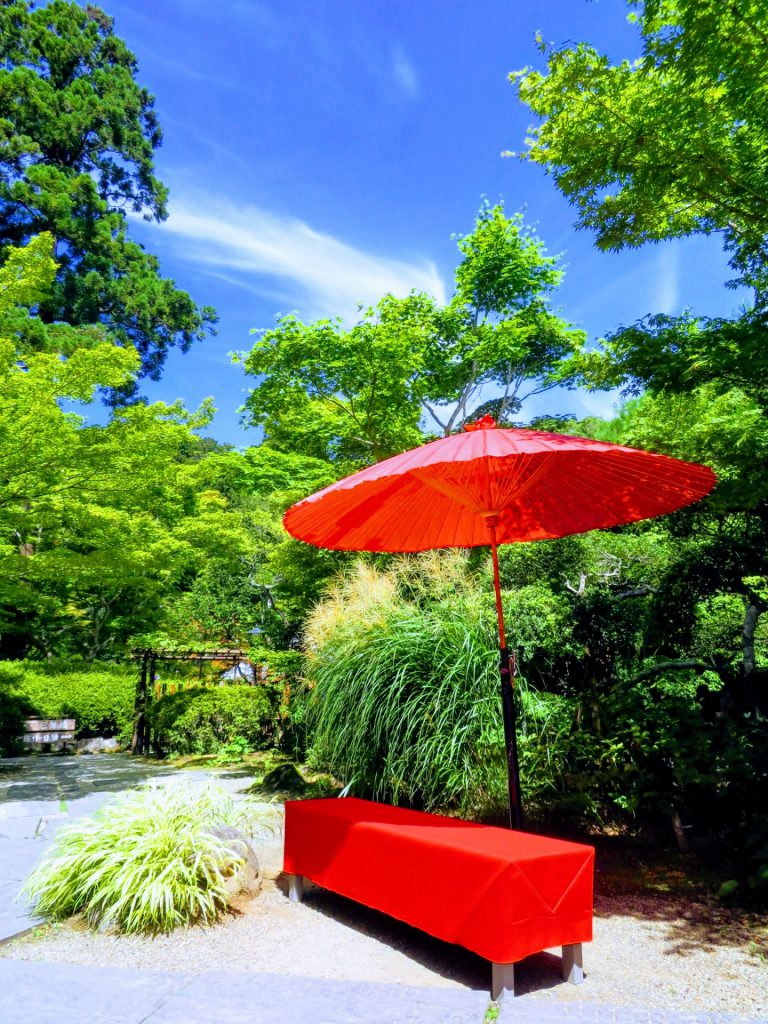 青空の下真っ赤な和傘が真っ赤な腰掛の横に立っていて緑に囲まれたお茶屋の雰囲気