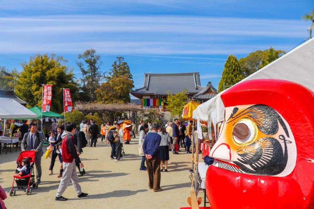 手前にだるまが置いてあり、奥に神社がある。神社へ向かう人々で賑わっている。