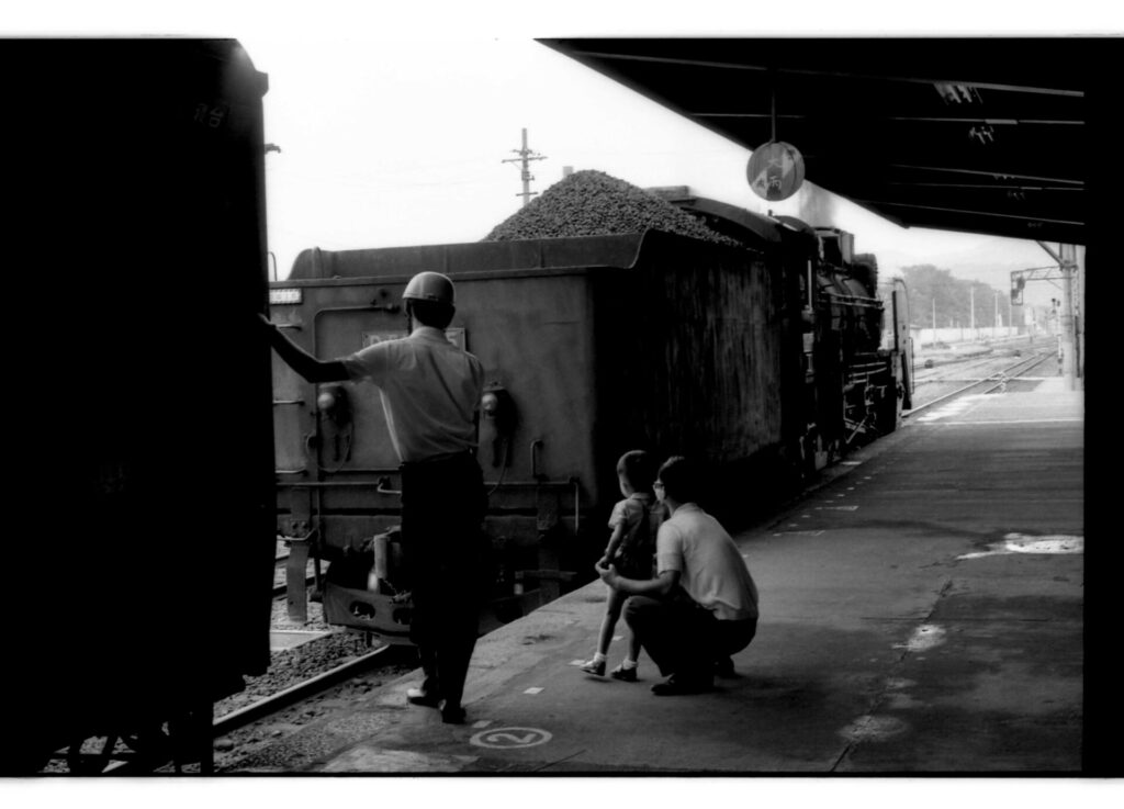 「D51895」の後ろに石炭が積んである。汽車の傍のホームに鉄道職員と、眺める父と子がいる