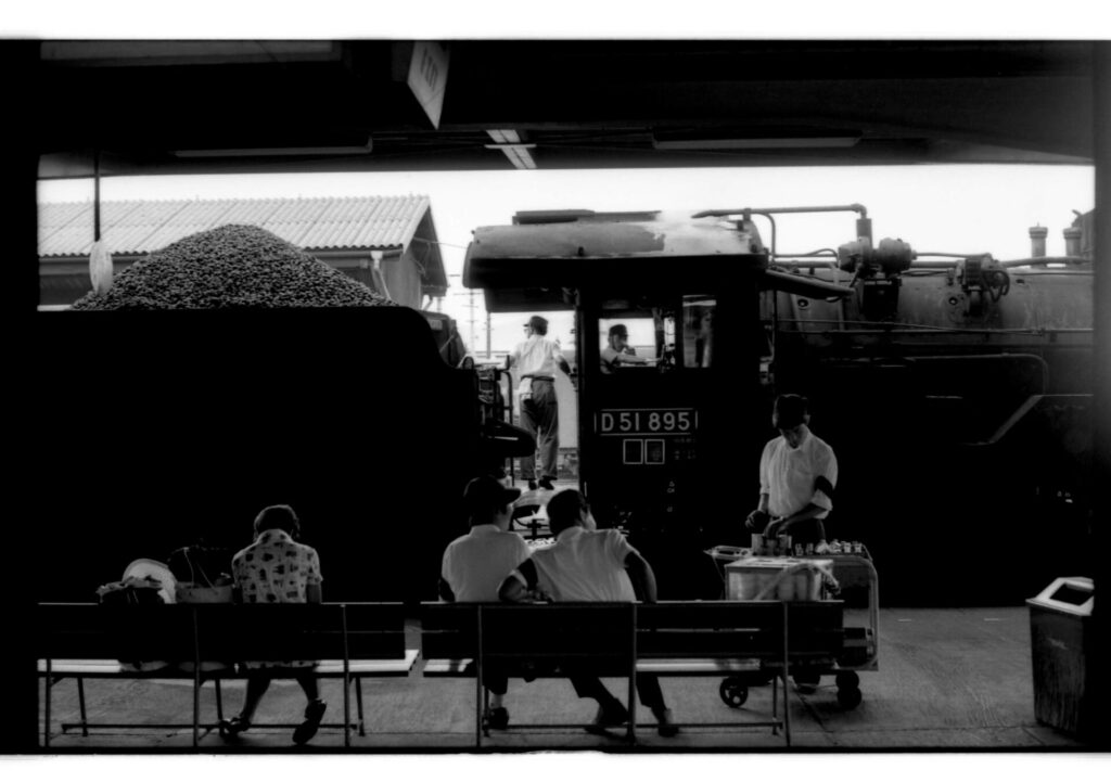駅のベンチに座る人々の目の前に石炭を満載した汽車「D51895」